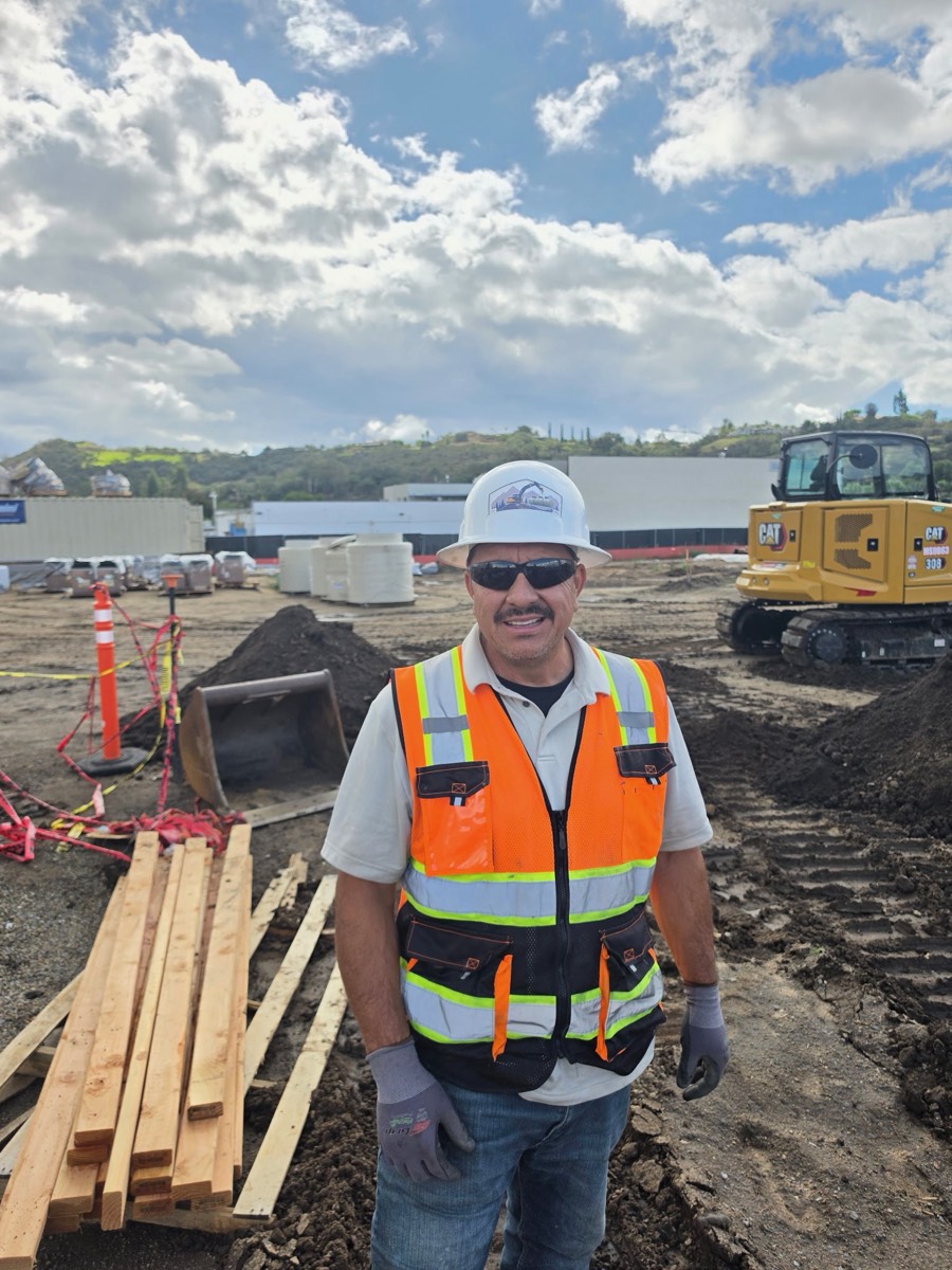 Frank Vasquez, owner of Vasquez Pipeline and Fuel Construction, on site in a hard hat with the company logo, mini excavator behind