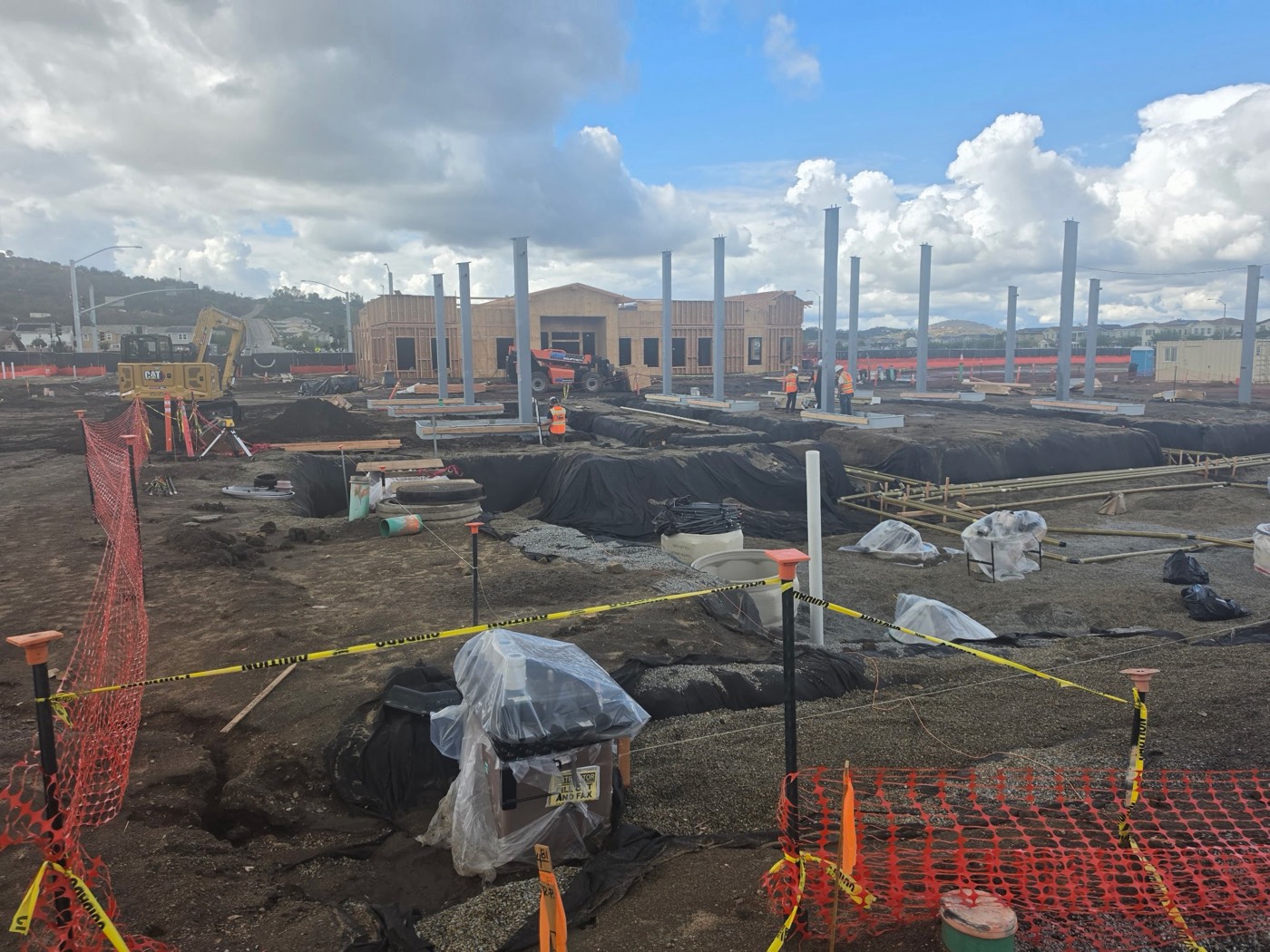 Fueling station under construction: canopy steel columns standing over lined trenches, with an excavator and the framed station building behind